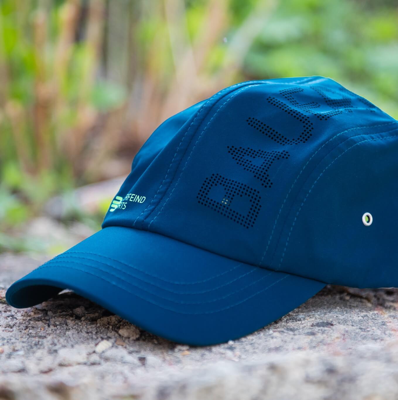 Blaue Bauerfeind Sports Cap in seitlicher Ansicht auf einem Stein, mit gebogenem Schirm, grünem Markenlogo und perforierter BAUERFEIND-Schrift auf dem Cap-Panel – ideal für frische Outdoor-Momente.