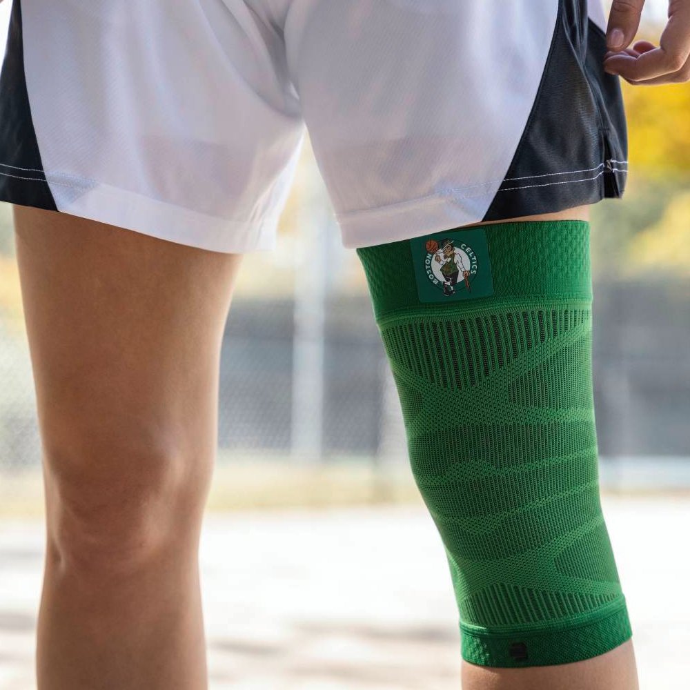 Nahaufnahme eines Spielers mit einer grünen Boston Celtics-Kniebandage auf einem Basketballplatz. Die Bandage zeigt das Teamlogo und passt sich ergonomisch an das Knie an.