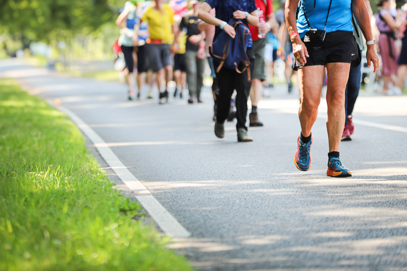 Wandergruppe beim 4Daagse Marsch Eine Gruppe von Wanderern läuft auf einer abgesperrten Straße im Grünen bei sonnigem Wetter. Im Vordergrund ist ein Mann mit blauer Kleidung und Sportschuhen zu sehen.
