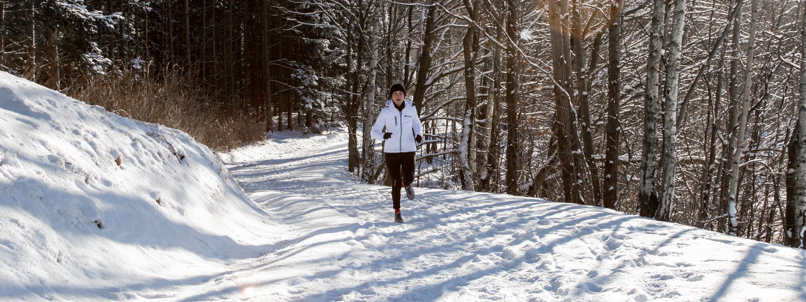 Frau in weißer Laufjacke joggt auf einem schneebedeckten Weg zwischen Bäumen und sieht in Richtung Kamera