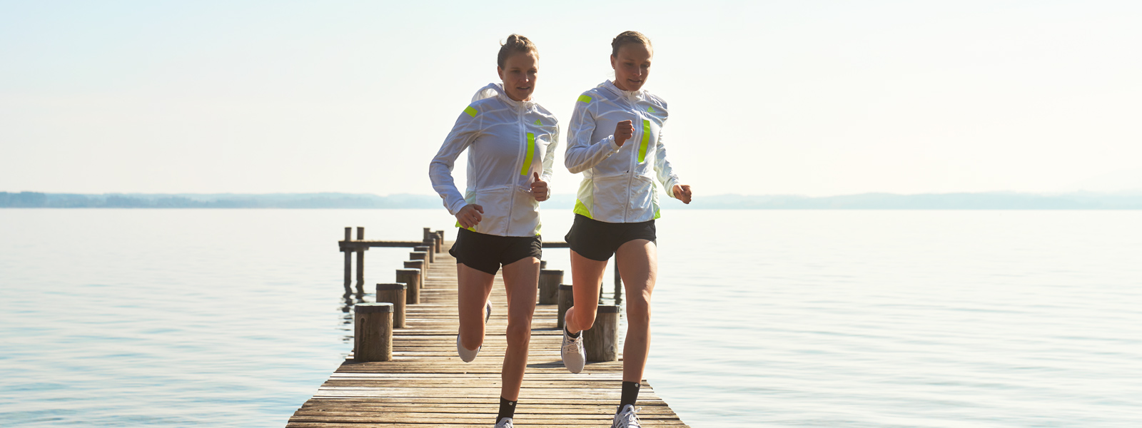 Wenn die Patellasehne streikt: Was tun bei einem Patellaspitzensyndrom? Zwei Läuferinnen in weißen Laufjacken und mit kurzen Hosen laufen über einen Steg in Richtung Ufer