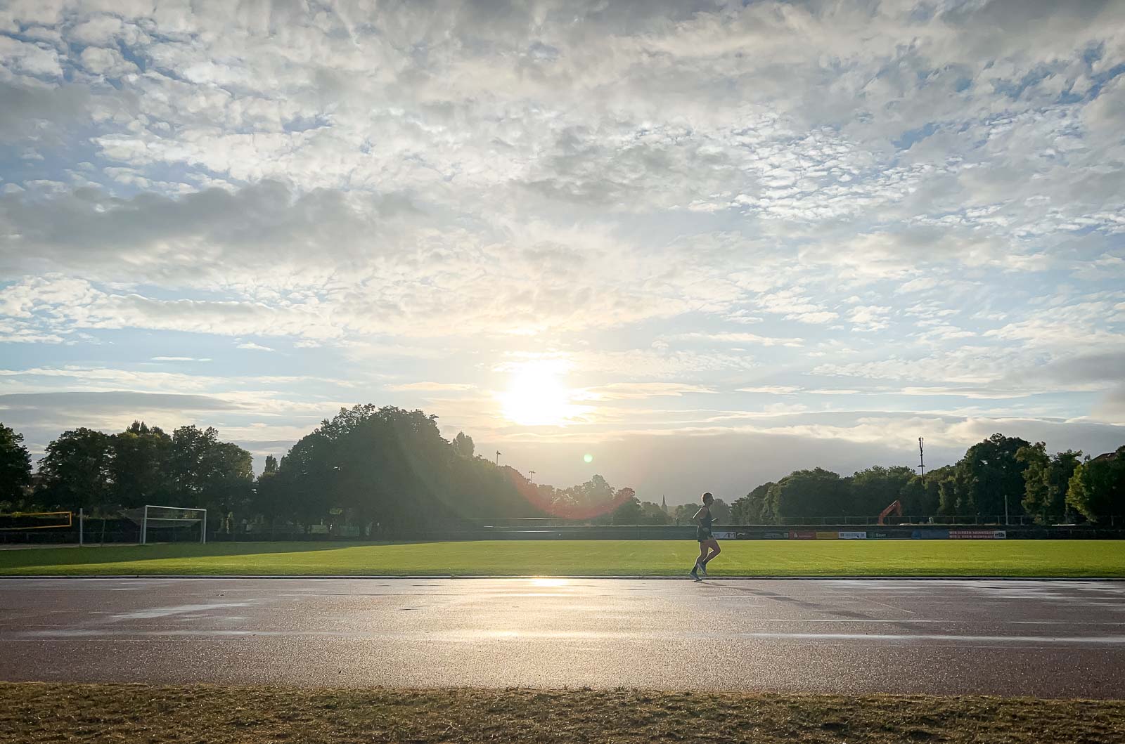 Läufer auf der Rennbahn – Training bei Sonnenaufgang für Ausdauer und Geschwindigkeit. Ein Läufer auf einer nassen Rennbahn bei Sonnenaufgang. Der Himmel ist bewölkt, aber die Sonne bricht durch, während der Läufer seine Runde beendet.