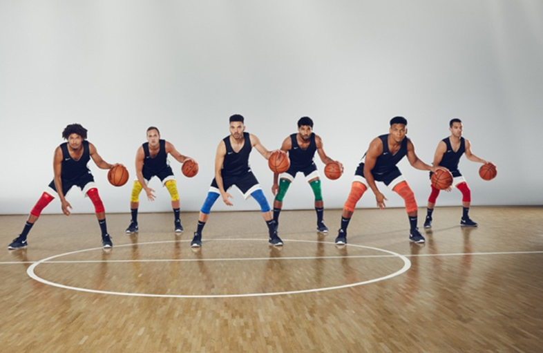 Sechs Basketballspieler stehen in einer Halle auf dem Spielfeld in leicht gebückter Position, jeder mit einem Basketball in der Hand. Sie tragen schwarze Trikots, weiße Shorts und Kompressionsbandagen.