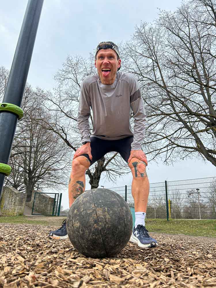 Ein Sportler steht nach einer intensiven Einheit im Outdoor-Trainingspark mit den Händen auf den Knien über einem Medizinball. Die Szene vermittelt Erschöpfung nach einem funktionellen Workout, wie es typisch für HYROX-Training ohne Gym ist.