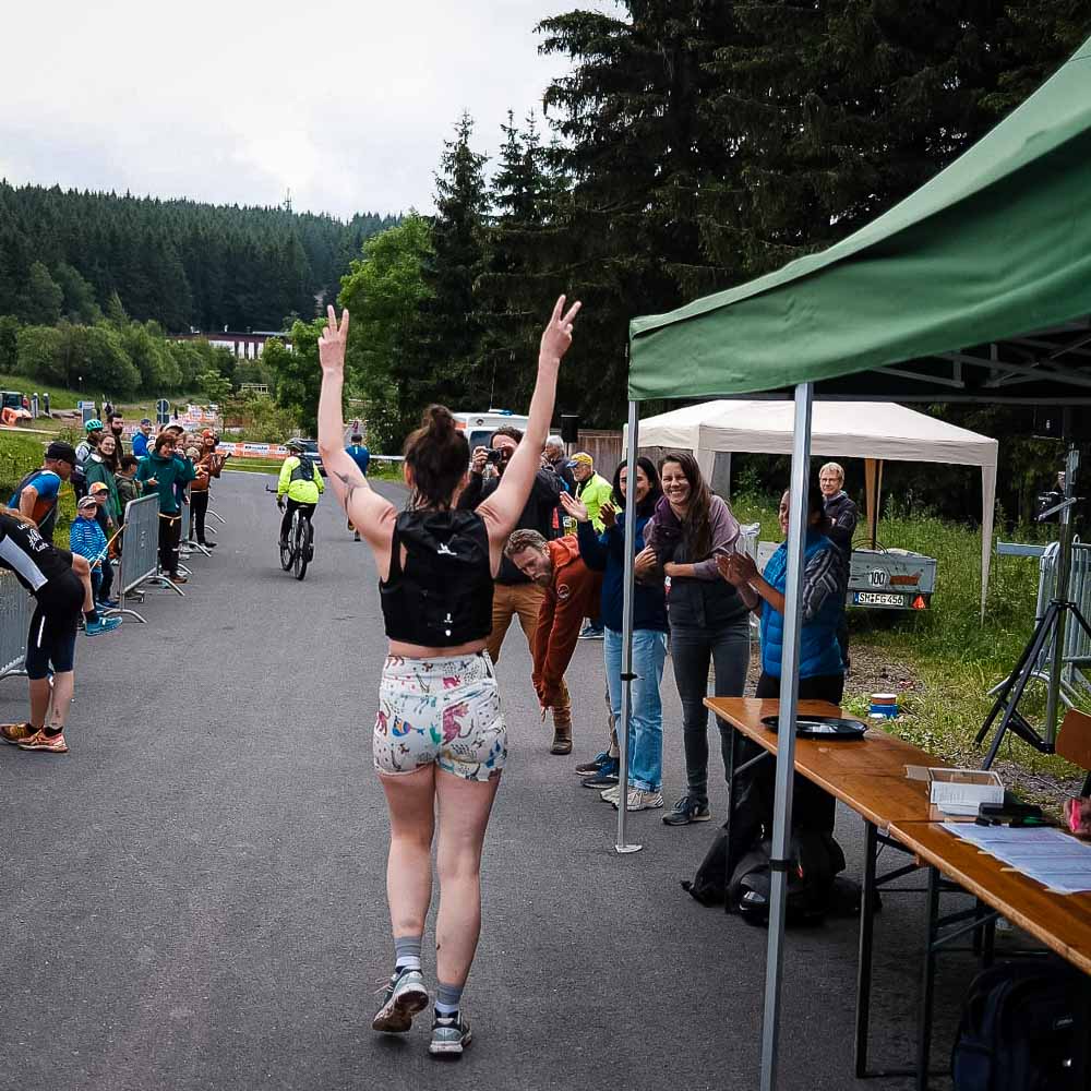 Den Rennsteig mit viel Teamunterstützung und guter Laune entlang joggen Eine Frau in Sportbekleidung joggt durch den Rennsteig und hebt dabei beide Arme, um das Peace-Zeichen zu machen, während die Zuschauer ihr zujubeln