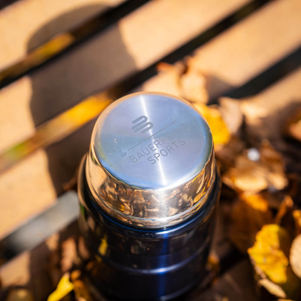 Elegantes Detail: Bauerfeind Sports Logo auf dem Thermosdeckel Nahaufnahme des glänzenden Deckels einer Thermoskanne, auf dem das Logo von Bauerfeind Sports eingraviert ist. Der Deckel liegt auf einer Holzbank, umgeben von herbstlichen Blättern.