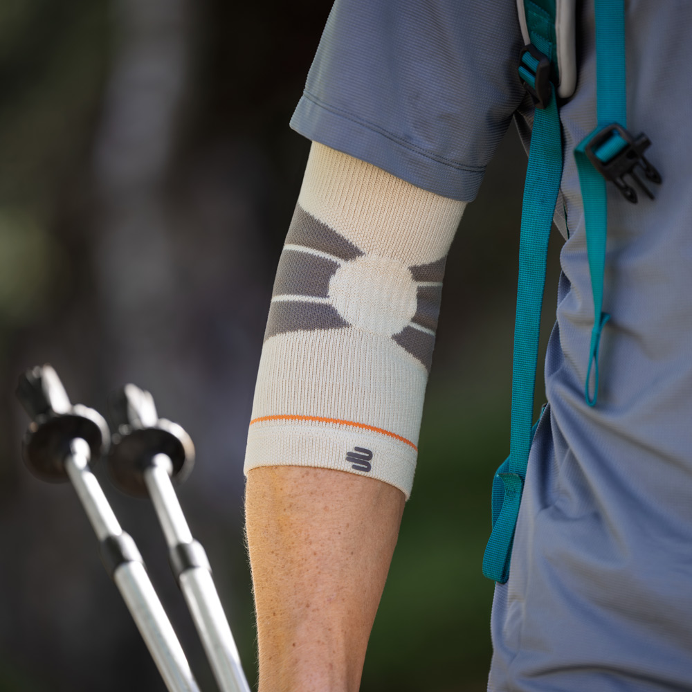 Ein Wanderer nutzt Trekkingstöcke auf einem schmalen Waldweg. Am rechten Arm trägt er eine Outdoor Compression Elbow Support Bandage in Sandstone-Farbe, die den Ellenbogen umschließt und unter dem kurzärmeligen Funktionsshirt sichtbar ist.