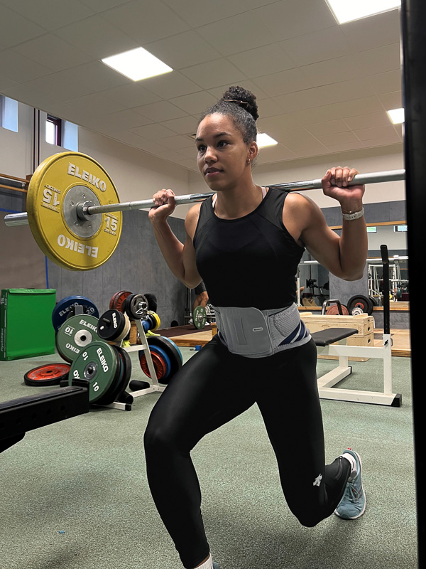 Deborah Levi nutzt eine Bauerfeind Bandage für Kraft und Stabilität. Deborah Levi macht im Fitnessstudio einen Ausfallschritt nach vorn. Auf ihren Schultern trägt sie eine Langhantel, um das Becken hat sie eine Bandage.