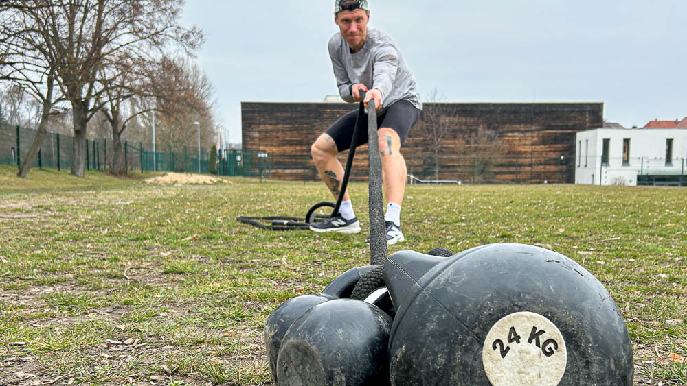 Athlet zieht beim Hyrox Training im Freien zwei Kettlebells mit einem Gurt über eine Wiese – funktionelles Krafttraining ohne Gym.