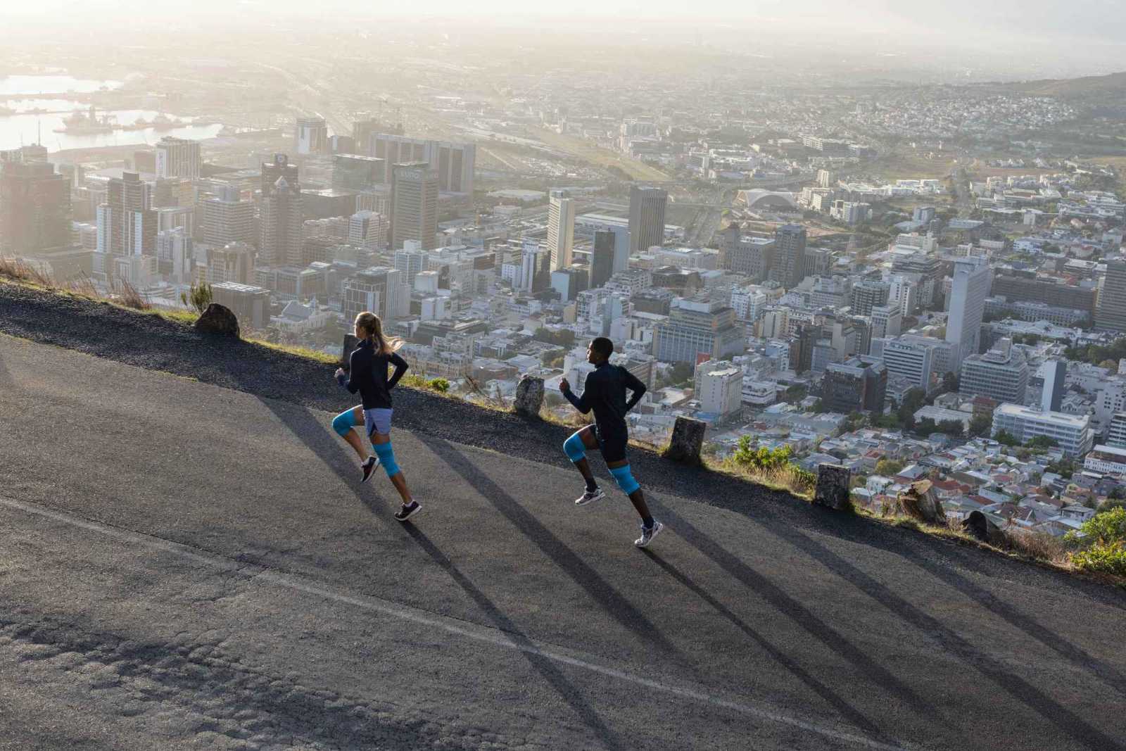 Ganz entspannt oberhalb der Metropole dank der Bauerfeind Sports Compression Knee Support Eine Frau und ein Mann mit blauen Kniebandagen laufen eine Straße oberhalb einer Großstadt hinauf