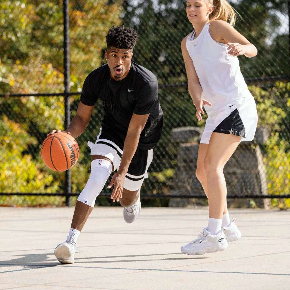 Zwei Basketballspieler im Eins-gegen-eins-Duell auf einem Outdoor-Court. Der Spieler in Schwarz trägt eine weiße Kompressions-Kniebandage und dribbelt den Ball, während der Gegenspieler in Weiß verteidigt.