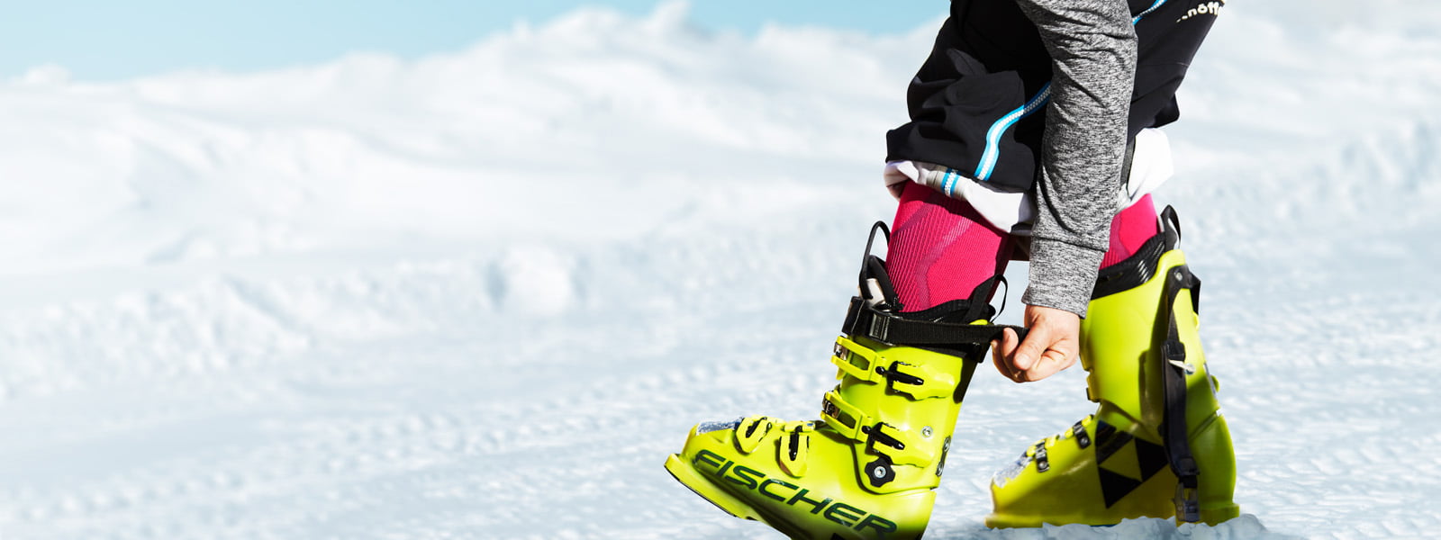 Skifahrer mit rosa Skisocken und grauem Ärmel zieht den Gurt bei seinem gelben Skischuh fest	 im Hintergrund Schnee
