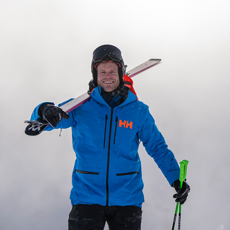 Bereit für die Piste Ein Mann steht vor einem nebligen Hintergrund, trägt eine blaue Jacke und einen Helm mit Skibrille und hat seine Skier auf der Schulter.