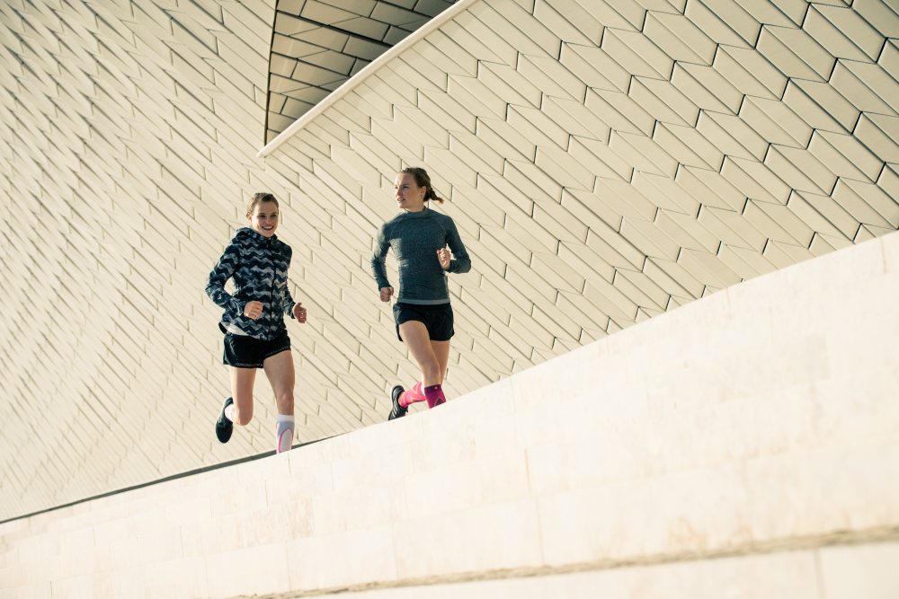 Zwei Läuferinnen laufen neben einer modern gestalteten Mauer im urbanen Umfeld und unterhalten sich