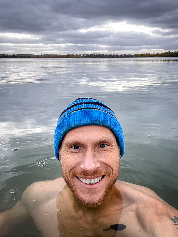 Lächeln trotz Gänsehaut – der ultimative Sprung ins kalte Nass! Ein Mann mit einer blauen Mütze lächelt in die Kamera, während er in einem kalten See steht. Der Himmel ist bewölkt, und das ruhige Wasser spiegelt die grauen Wolken wider.