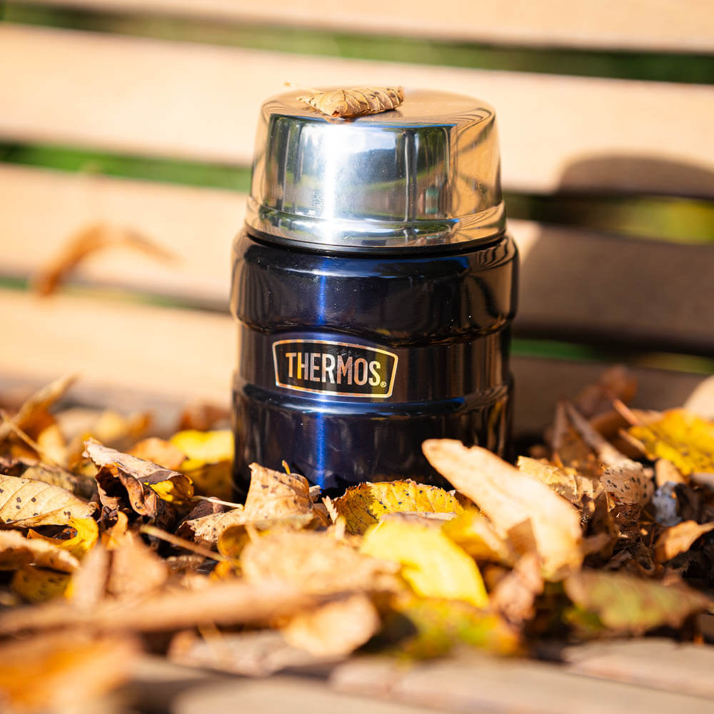 Herbstliche Pause: Mehr Wärme durch unsere Thermoskanne Eine blaue Thermoskanne steht auf einer Holzbank und ist von Herbstblättern umgeben. Auf dem glänzenden Deckel liegt ein kleines Blatt. Die Kanne trägt den Schriftzug "Thermos" und strahlt in der herbstlichen Umgebung.