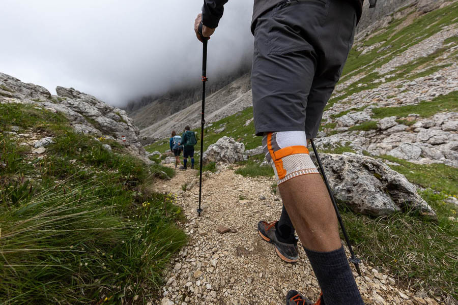 Ein Wanderer mit Trekkingstöcken und einer Kniebandage geht auf einem felsigen Pfad durch eine bergige Landschaft. Im Hintergrund sind weitere Wanderer und Wolken über den Bergen zu sehen.