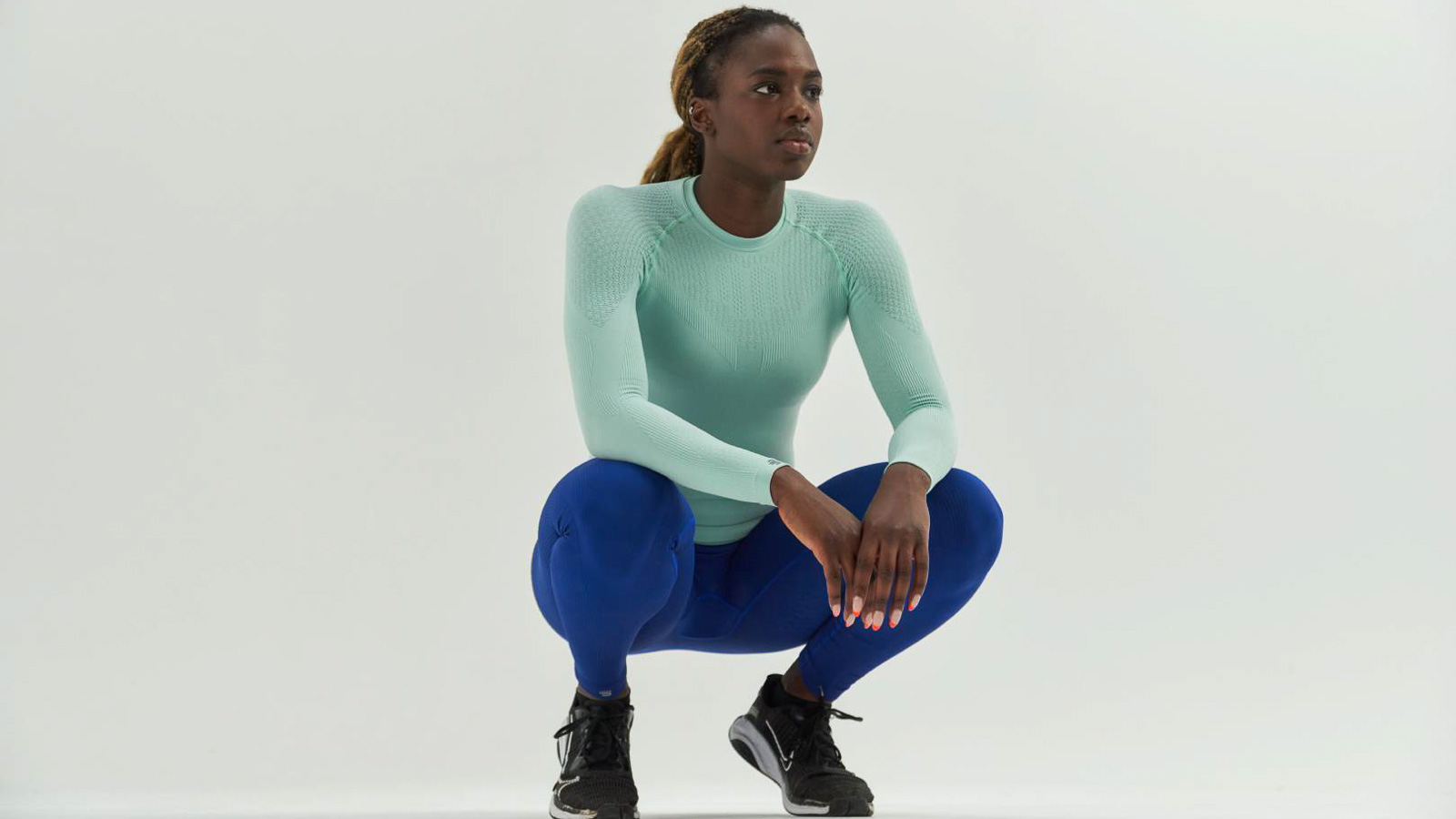 Athletin in mintgrünem Kompressions-Shirt und blauer Kompressionshose in Hockstellung vor weißem Hintergrund.
