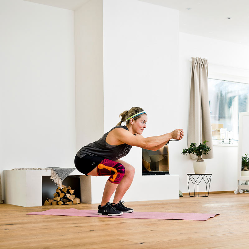 Skigymnastik - die Abfahrtshocke mit der Sports Knee Support Frau mit Kniebandage simuliert die Ski-Abfahrtsposition in einem Kaminzimmer	 Rücken gerade	 Arme mit ineinander verschränkten Händen nach vorn