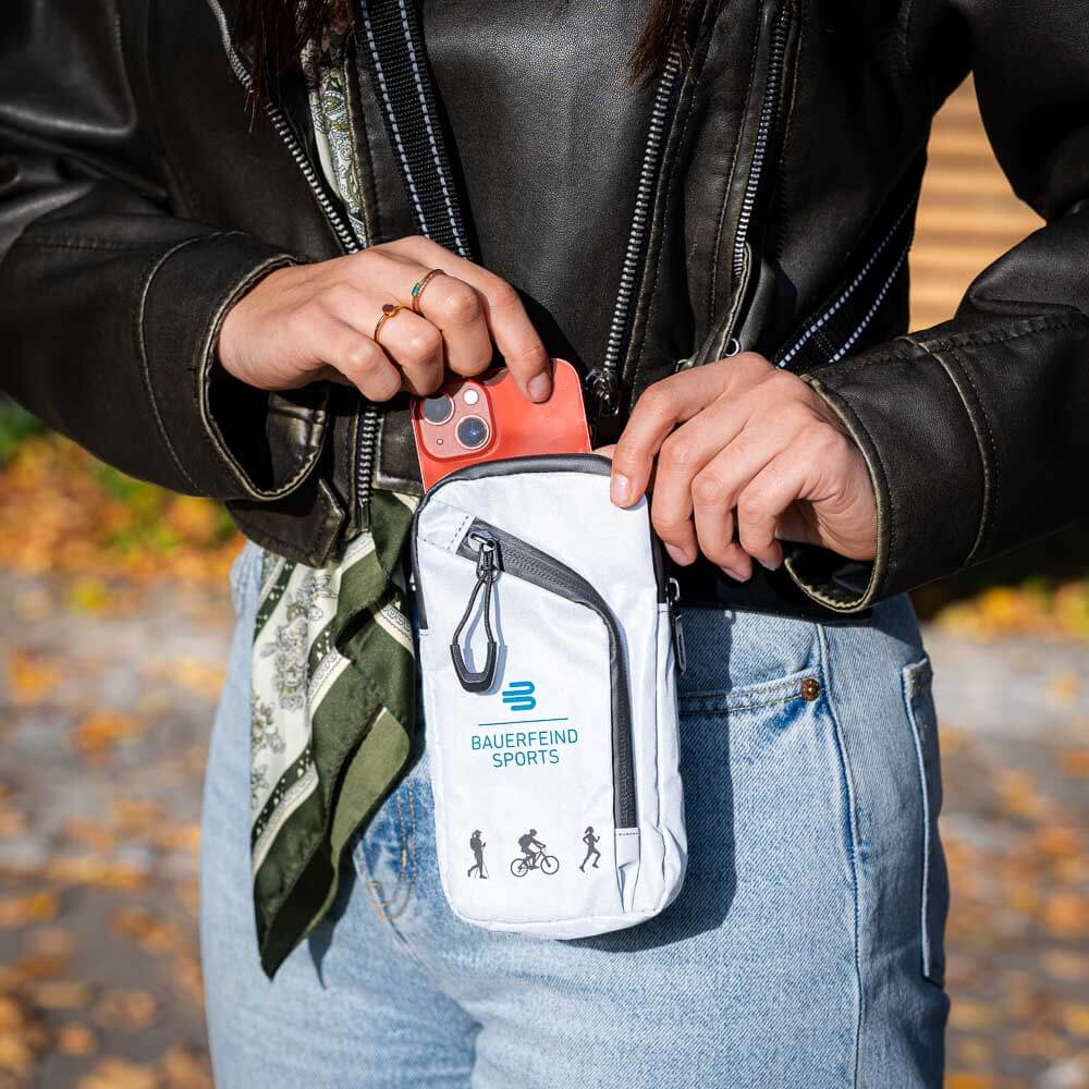 Kompakt und funktional: Die perfekte Tasche für unterwegs Eine Person verstaut ein Smartphone in der grauen Bauerfeind Sports Tasche, die an der Hüfte getragen wird. Die Tasche ist kompakt und eignet sich perfekt für Outdoor-Aktivitäten.