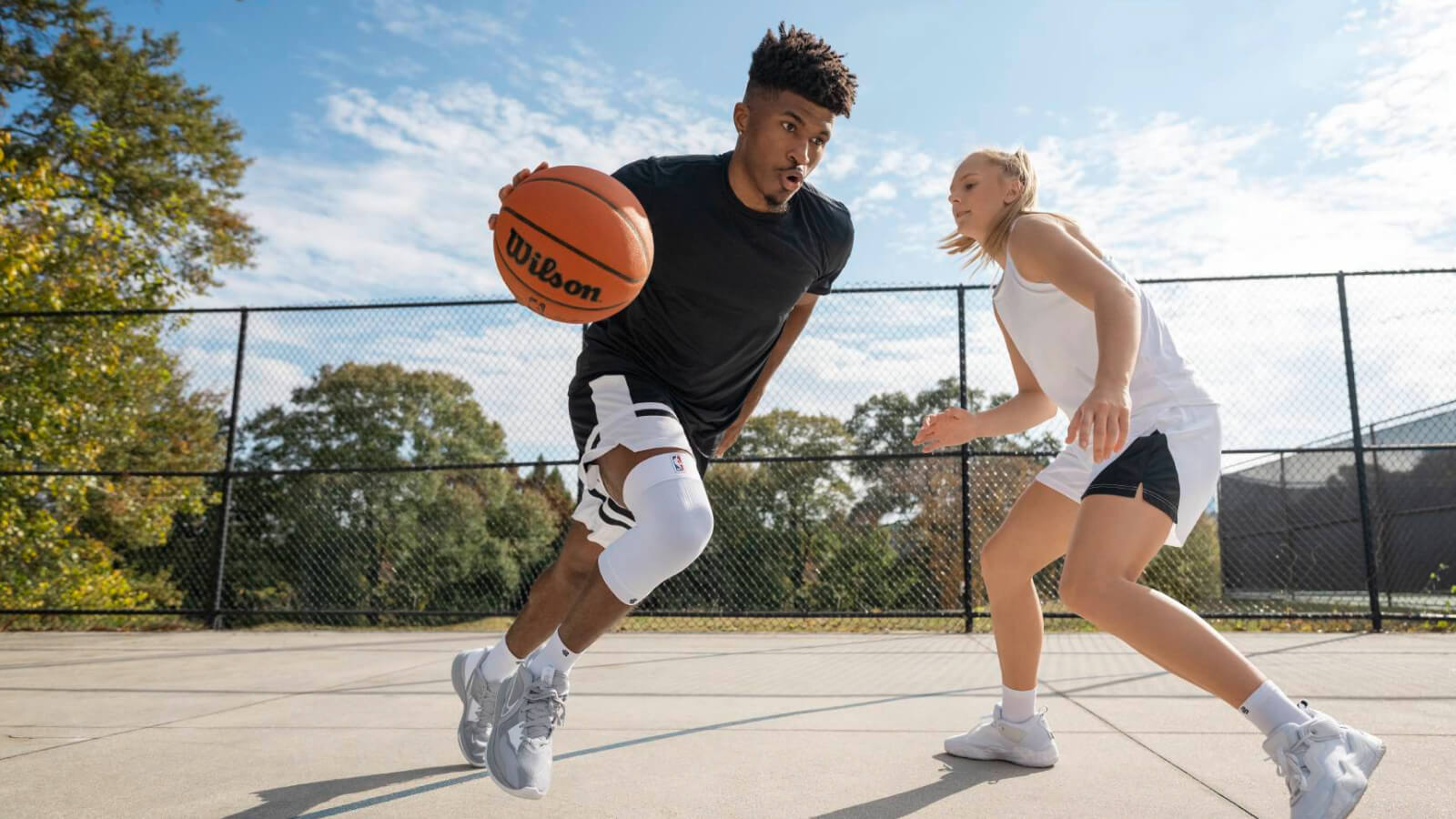 Ein Basketballspieler treibt den Ball energisch über einen Freiplatz, verfolgt von einer Gegnerin. Er trägt eine weiße Kniebandage, die seinen Bewegungsapparat unterstützt.