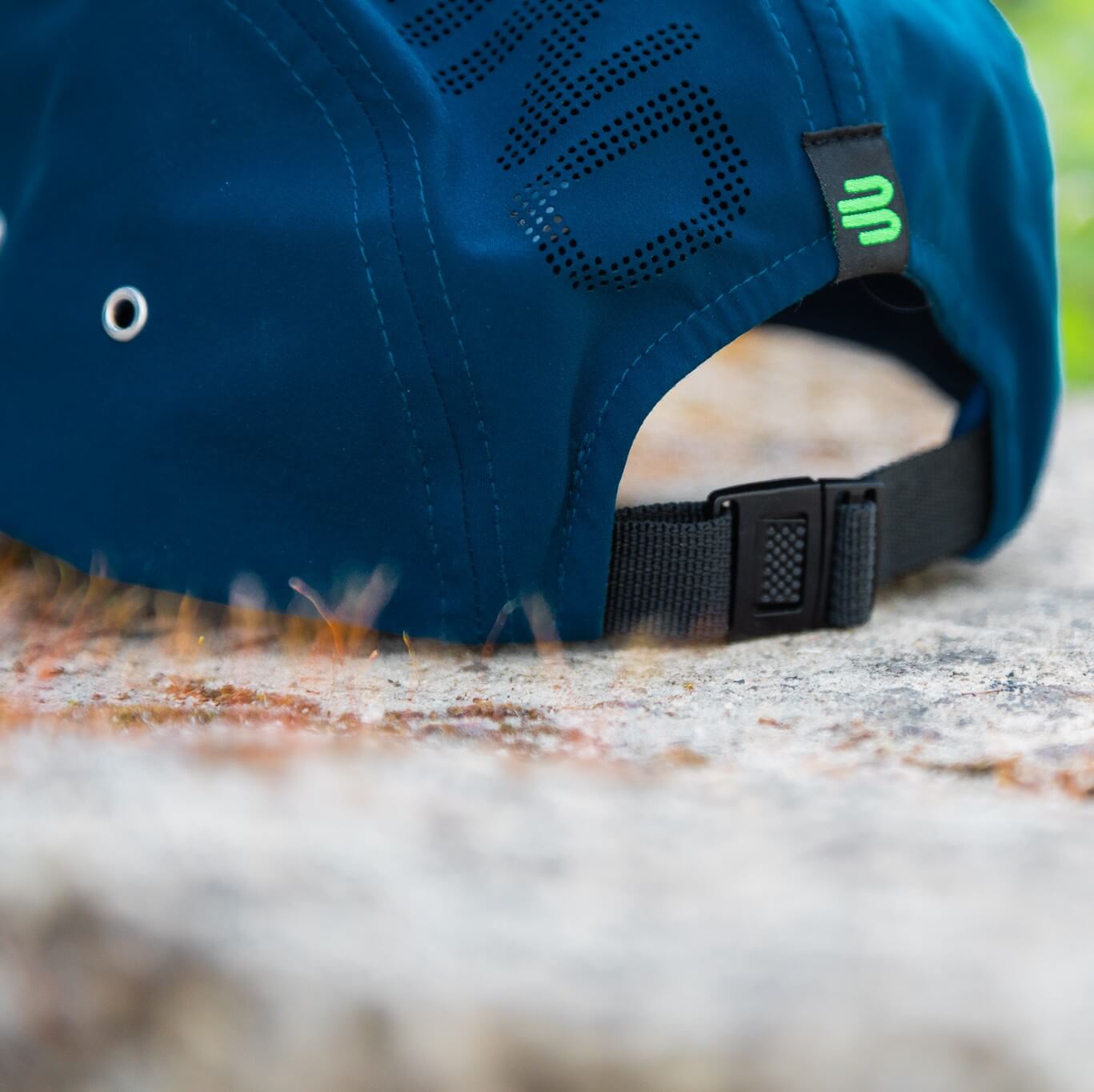 Nahaufnahme der Rückseite einer blauen Bauerfeind Sports Cap mit schwarzem verstellbarem Verschluss, grünem Logo-Label, seitlicher Metallöse und perforierter Schrift – praktische Details für sicheren Halt bei aktiven Outdoor-Momenten.