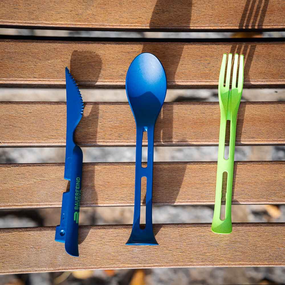 Das komplette Set: Messer, Gabel und Löffel für unterwegs Ein auseinandergelegtes Besteck-Set mit einem blauen Messer, einem blauen Löffel und einer grünen Gabel liegt nebeneinander auf einer Holzbank.
