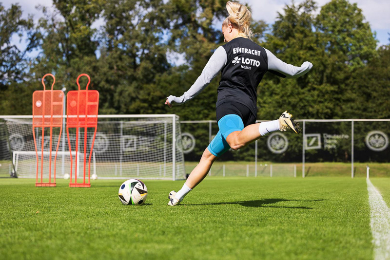 Spielerin von Eintracht Frankfurt beim Torschusstraining Eine Spielerin von Eintracht Frankfurt schießt mit voller Kraft den Ball auf ein Tor im Hintergrund. Sie trägt ein Trainingsoutfit mit Eintracht-Logo und eine blaue Kniebandage von Bauerfeind Sports.
