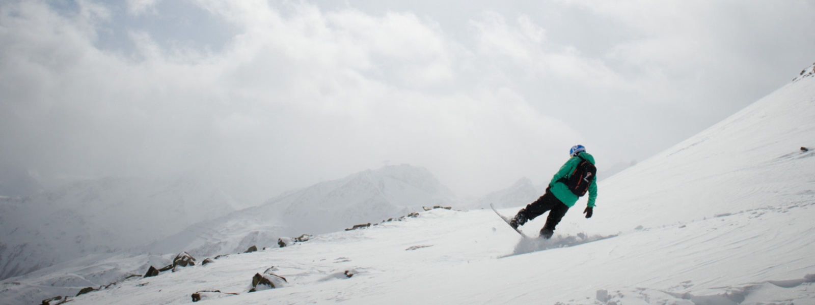 Snowboarder in grüner Jacke bremst mit dem hinteren Teil seines Boards vor einer Kuppe	 im Hintergrund sieht man verschneite Berggipfel durch die Wolken