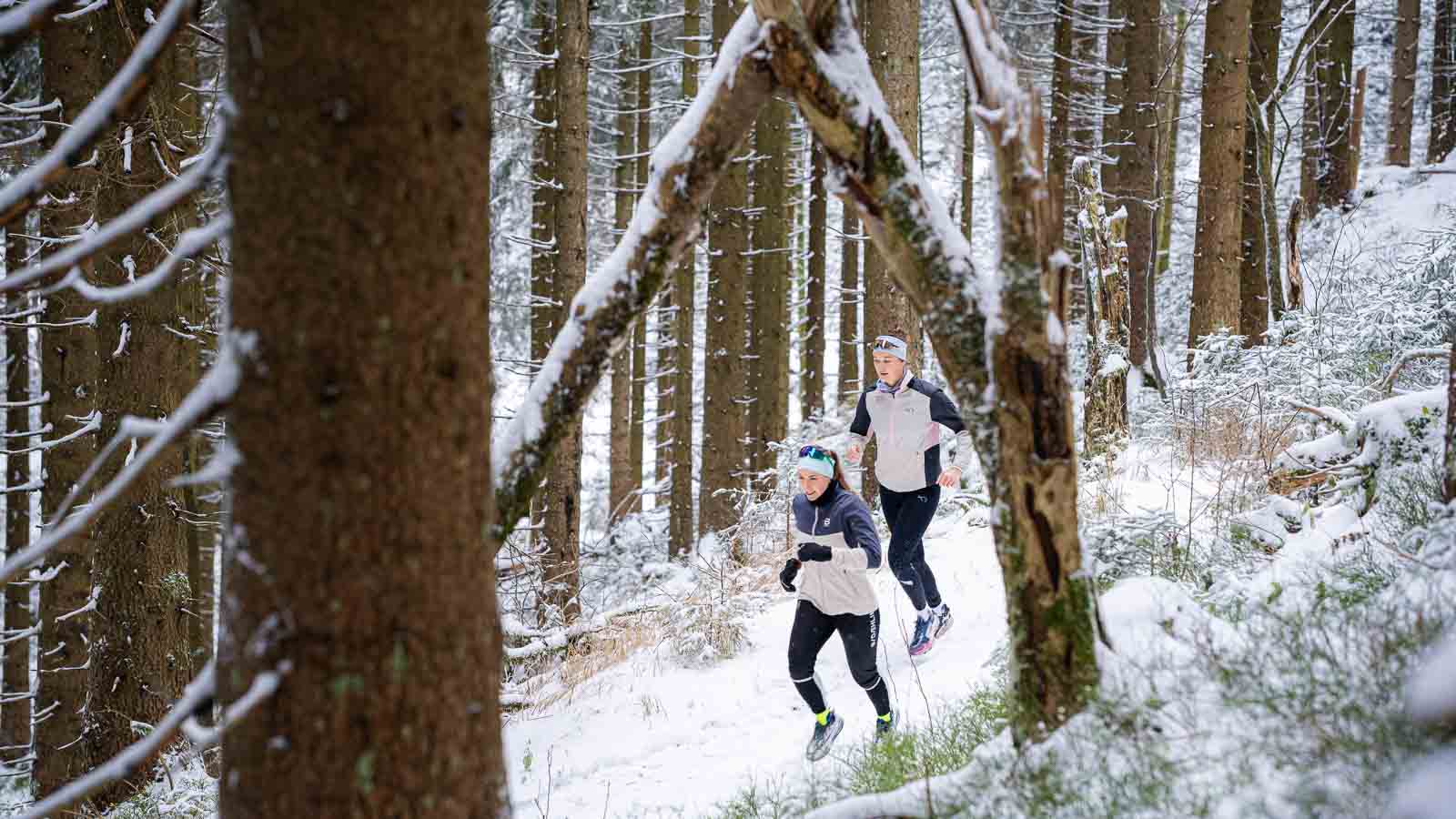 Zwei Personen laufen auf einem verschneiten Waldweg zwischen hohen, mit Schnee bedeckten Bäumen. Beide tragen Winterlaufsportbekleidung, darunter Laufjacken, Leggings und Handschuhe.