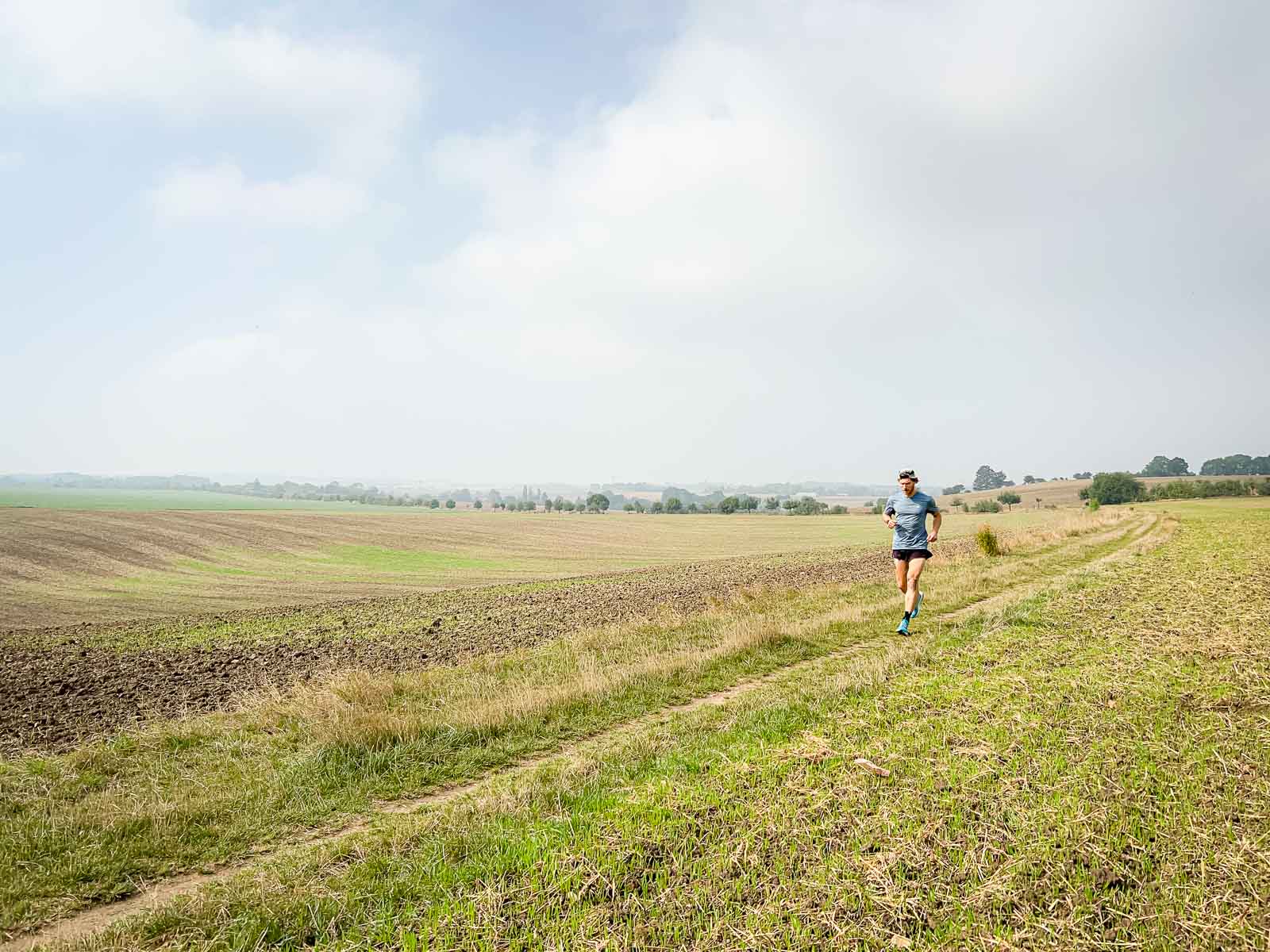 Lauftraining auf einem Feldweg – Weitläufige Landschaft für Ausdauerläufe. Ein Läufer läuft auf einem Feldweg in einer ländlichen Landschaft bei leicht bewölktem Himmel. Die Umgebung ist ruhig und weitläufig, perfekt für Ausdauertraining.
