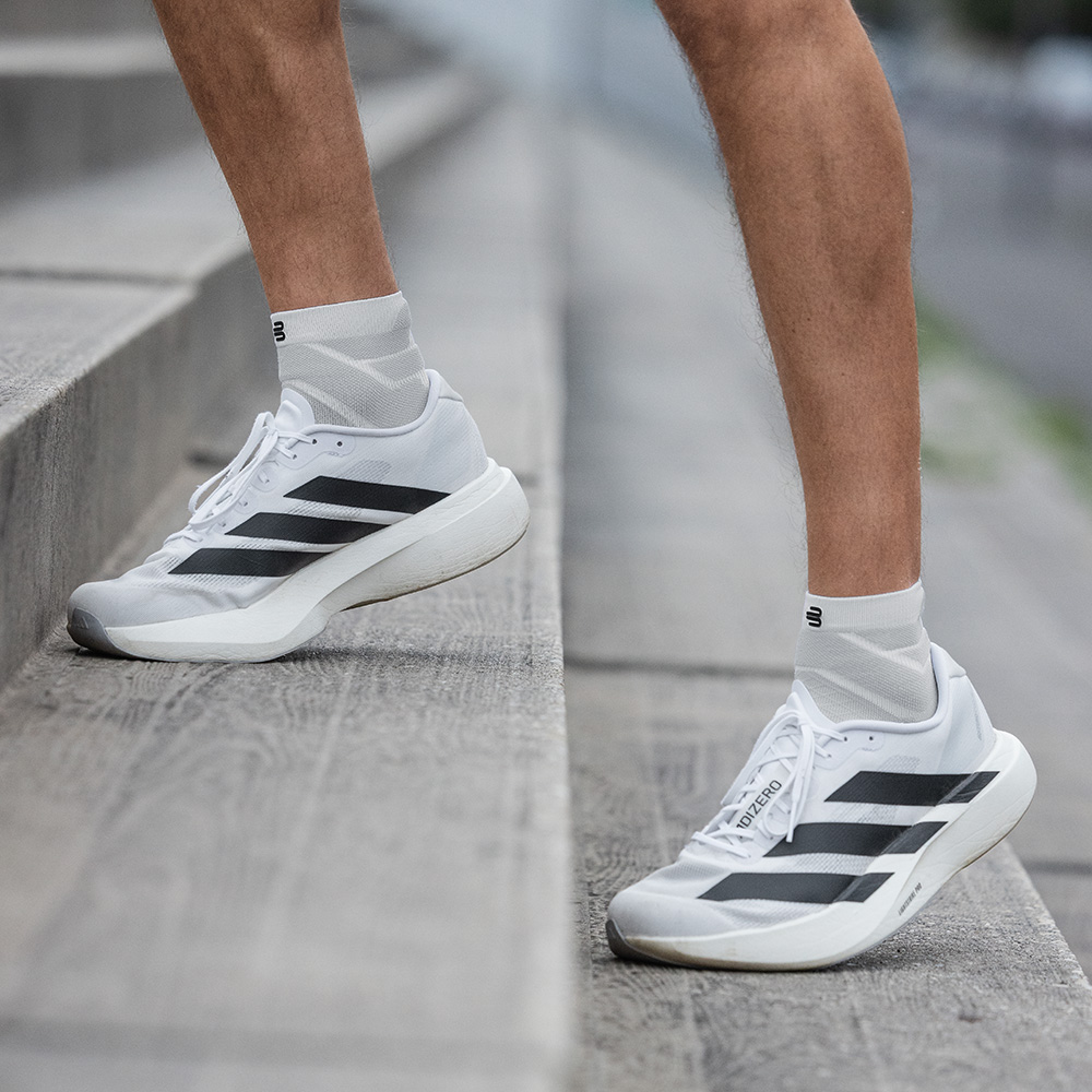 Ein Mann steht mit leicht angewinkelten Beinen auf einer Treppe. Zu sehen sind seine weißen, kurzen Run Performance Socks und weiße Laufschuhe mit schwarzen Streifen.