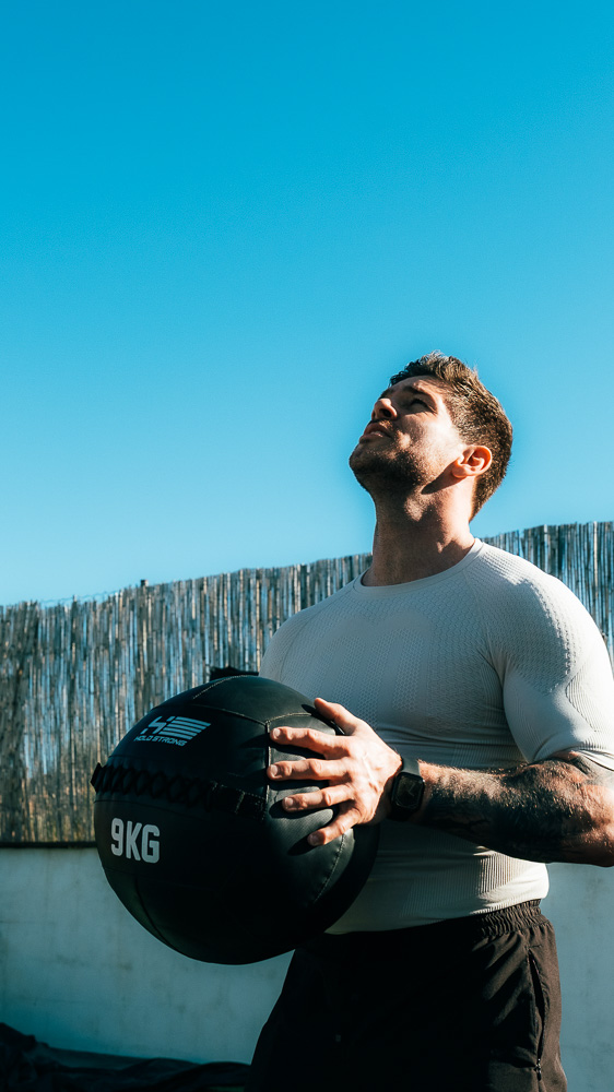 Athlet steht draußen mit einem schwarzen 9-kg-Medizinball vor dem Oberkörper, trägt ein helles Kompressionsshirt und schaut unter blauem Himmel nach oben – funktionelles Training für Hybrid-Athleten.
