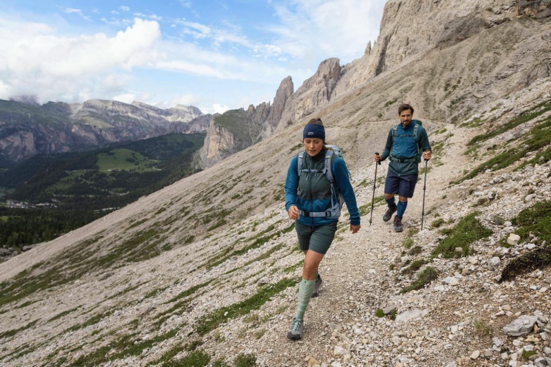 Eine Frau und ein Mann wandern auf einem Gebirgsweg umgeben von Felsen und tragen Rucksack	 Teleskopstöcke und lange Wandersocken