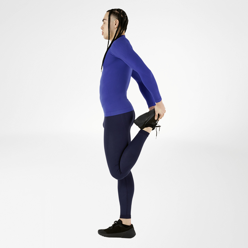 Ein Sportler in einem blauen Kompressionsoutfit macht eine Dehnübung und hält dabei ein Bein nach hinten, vor einem weißen Hintergrund. Das Outfit bietet hohen Tragekomfort und optimale Unterstützung der Muskulatur.