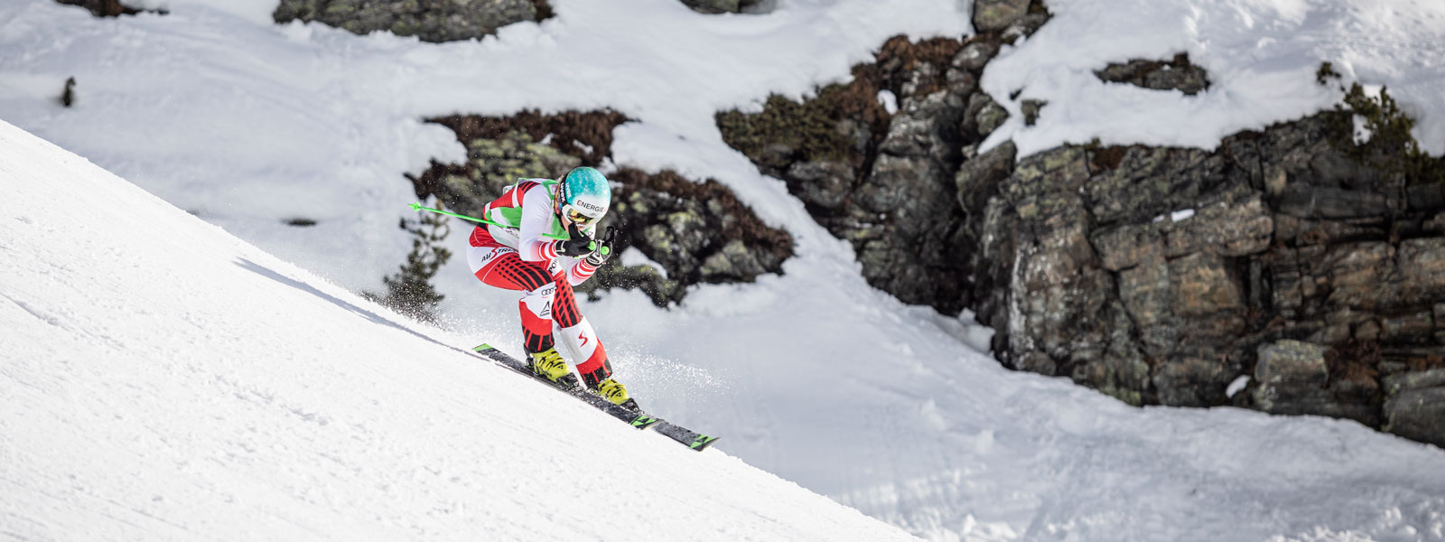 Skiläuferin im rot-weißen Rennanzug in Abfahrtsposition bei einem leichten Schwung mit großen	 schneebedeckten Felsen im Hintergrund