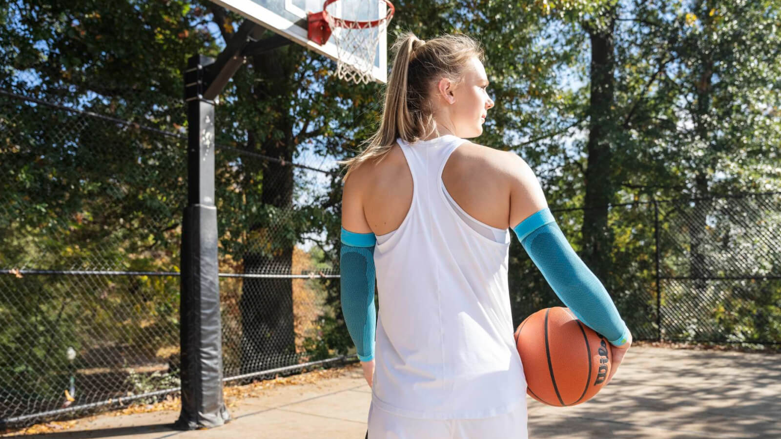 Bereit für den nächsten Wurf. Eine Basketballspielerin steht auf einem Outdoor-Platz mit einem Basketball in der rechten Hand. Sie trägt hellblaue Kompressions-Arm Sleeves und blickt konzentriert auf den Korb.