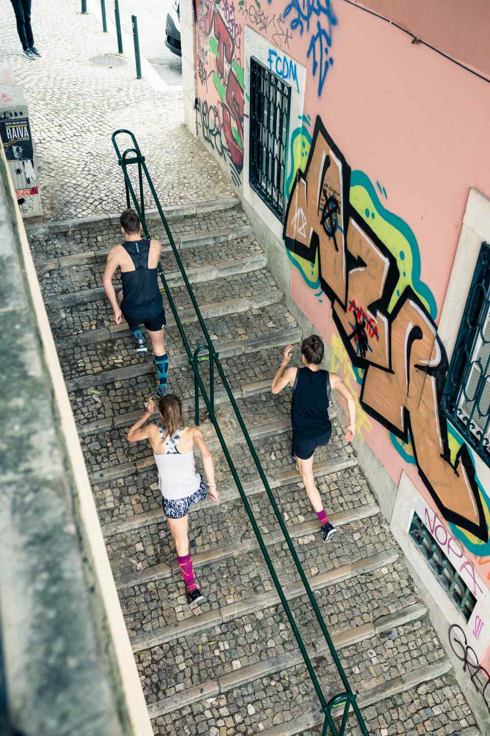 Vogelperspektive: Ein Mann und zwei Frauen in Laufoutfits laufen eine Treppe in einer Altstadt mit Graffitis an den Wänden nach oben