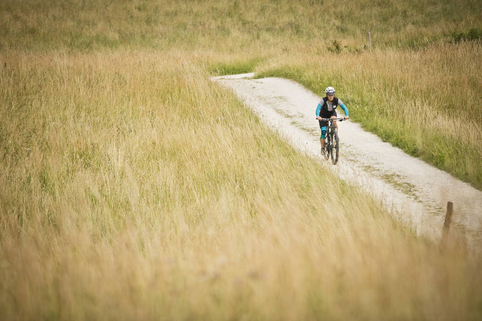 Frau mit Armsleeves und Kniebandage im Bildhintergrund fährt auf einem Mountainbike über einen Weg zwischen hohem Gras