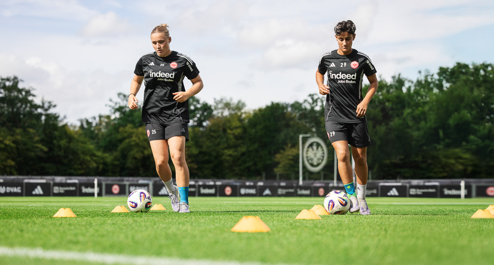 Zwei Spielerinnen von Eintracht Frankfurt trainieren auf dem Fußballplatz mit Koordinationshütchen und Ball. Sie tragen kurze schwarze Trainingsoutfits und sportliche Kompressionsprodukte von Bauerfeind Sports, ideal für dynamische Bewegungsabläufe.