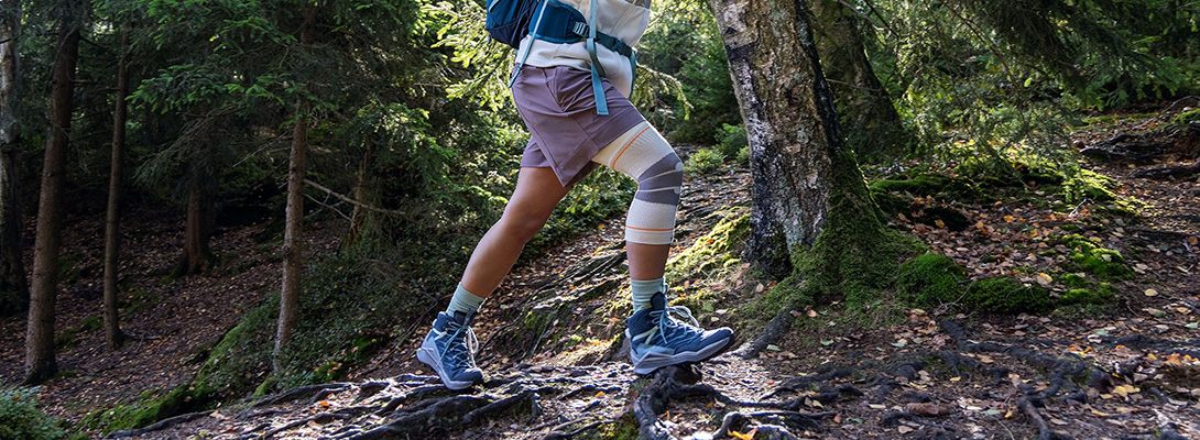 Person beim Wandern auf einem wurzeligen Waldpfad, trägt Wanderschuhe, Socken und eine Kniebandage am rechten Bein.
