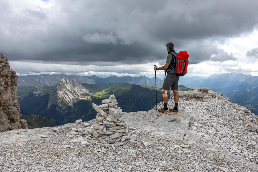 Wanderer auf einem Felsvorsprung mit Wander Kniebandage Eine Person mit einem roten Rucksack steht auf einem Felsvorsprung in den Dolomiten und blickt in die Ferne. Der Himmel ist bewölkt und es gibt eine Steinmännchen-Konstruktion im Vordergrund.