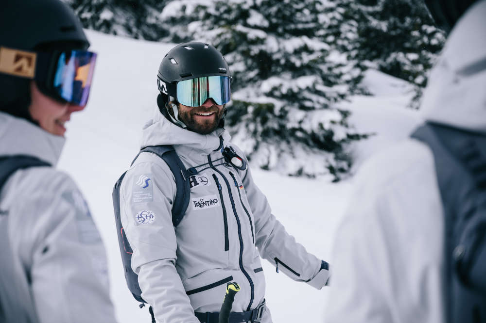 Gut gelaunter Skifahrer mit schwarzem Helm	 grauer Jacke und Rucksack	 Aufnahme zwischen zwei weiteren Skifahrern