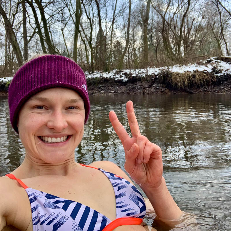 Anna Hahner beim Eisbaden im Fluss Anna Hahner steht lächelnd bis zur Brust im kalten Wasser, umgeben von schneebedeckter Landschaft und kahlen Bäumen, und zeigt ein Peace-Zeichen mit den Fingern.