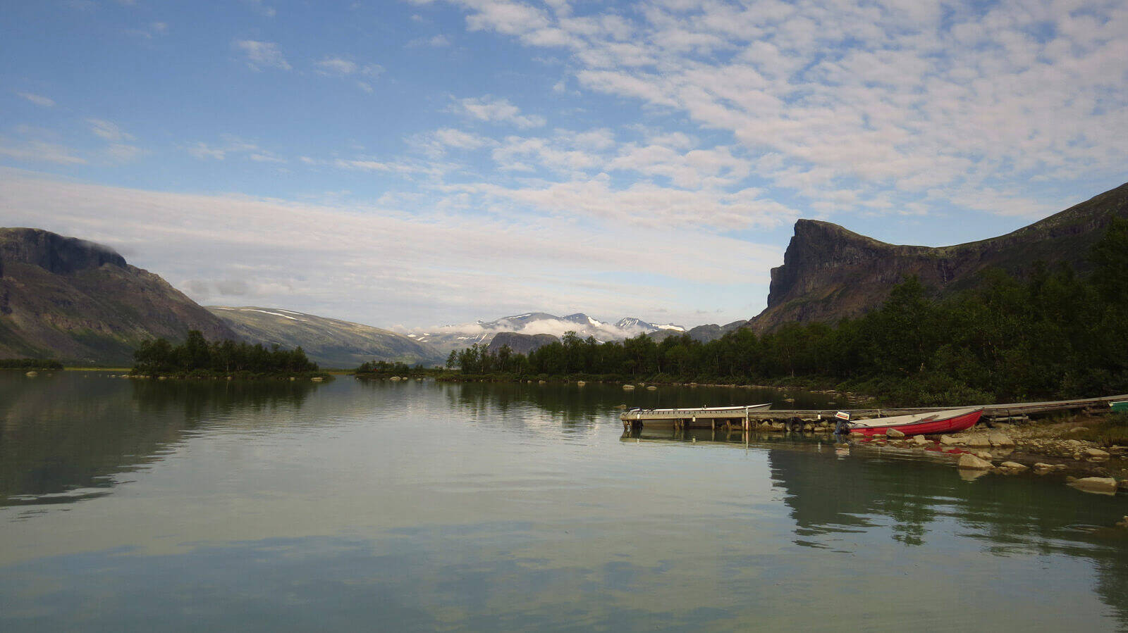 Der Kungsleden gilt nicht umsonst als einer der beliebtesten Wanderwege in Europa Panorama eines Sees umgeben von Bäumen und kleinen Bergen	 am rechten Rand liegt ein kleines Boot am Steg