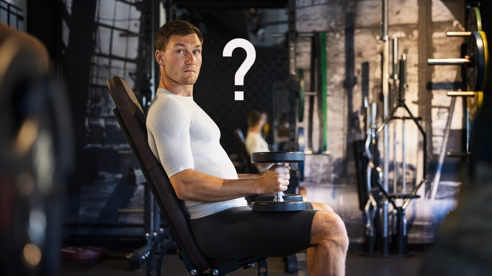 Ein Mann sitzt in einem Fitness-Studio auf einer Hantelbank. Er trägt ein weißes Kompressions-Shirt.