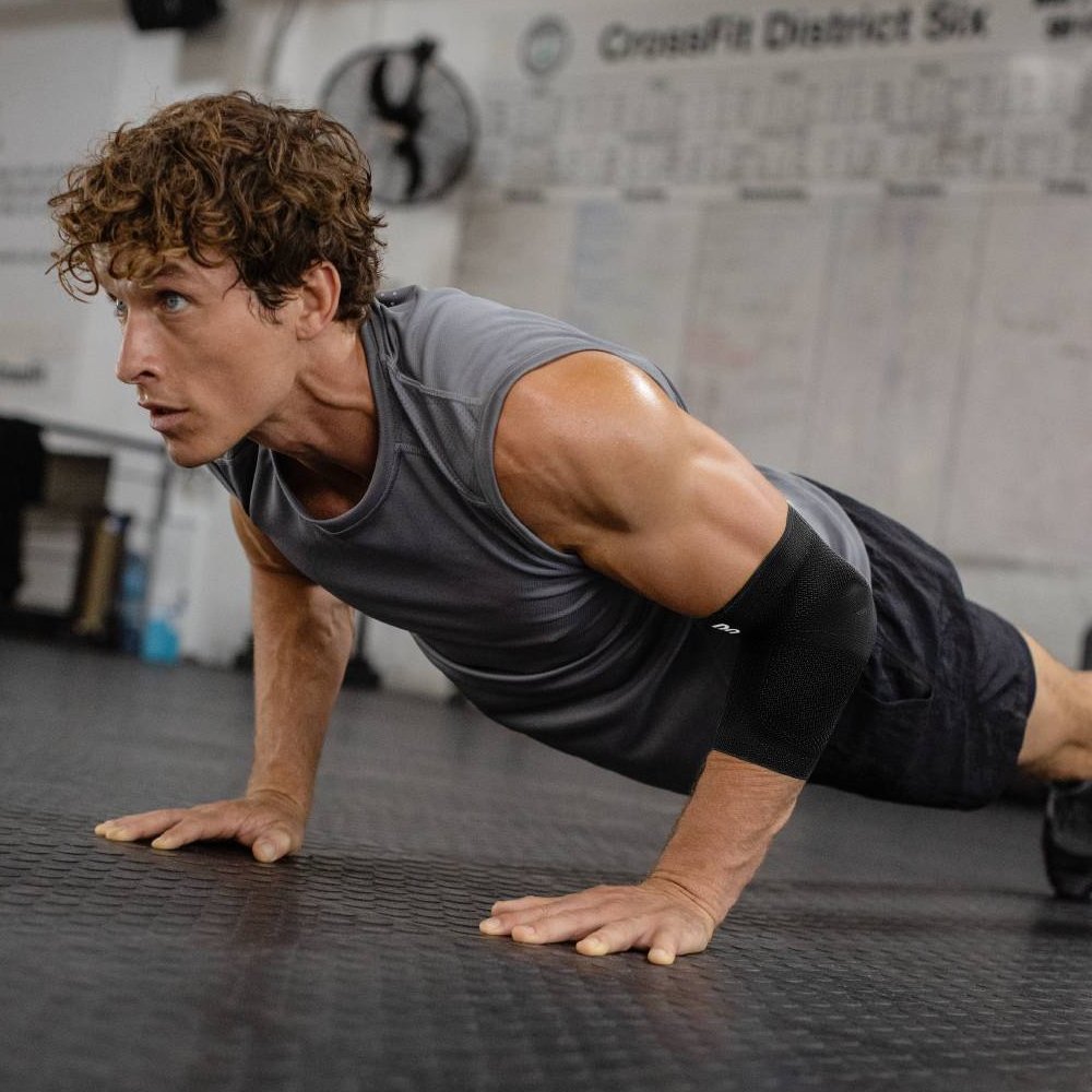 Ein Athlet im Fitnessstudio, der während eines intensiven Liegestütz-Trainings eine schwarze Ellenbogenbandage trägt. Die Bandage bietet stabilen Halt und optimalen Schutz für den Ellenbogen bei Krafttraining und sportlichen Herausforderungen.