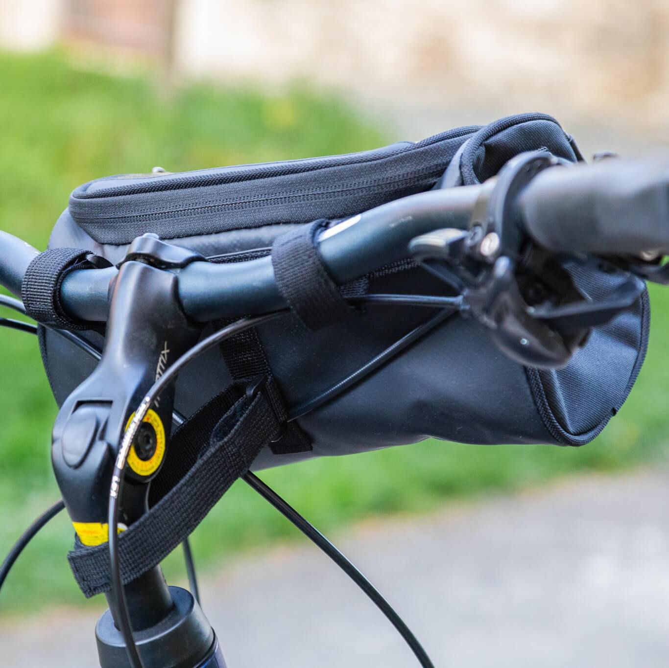 Nahaufnahme der schwarzen Bike Bag am Fahrradlenker, bei der Befestigungsriemen, Bremshebel, Kabelzüge und der obere Reißverschluss gut sichtbar sind – die Tasche sitzt kompakt am Cockpit und hält wichtige Dinge während der Fahrt griffbereit.