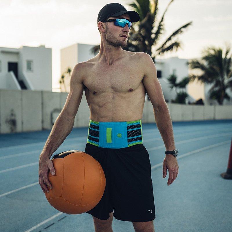 Starker Rücken, starkes Training – mit stabiler Unterstützung das Beste aus sich herausholen. Ein sportlicher Mann mit Sonnenbrille und Baseballkappe steht auf einer Laufbahn und trägt eine leuchtend blaue Rückenbandage mit grünen Elementen. In der Hand hält er einen großen Medizinball.