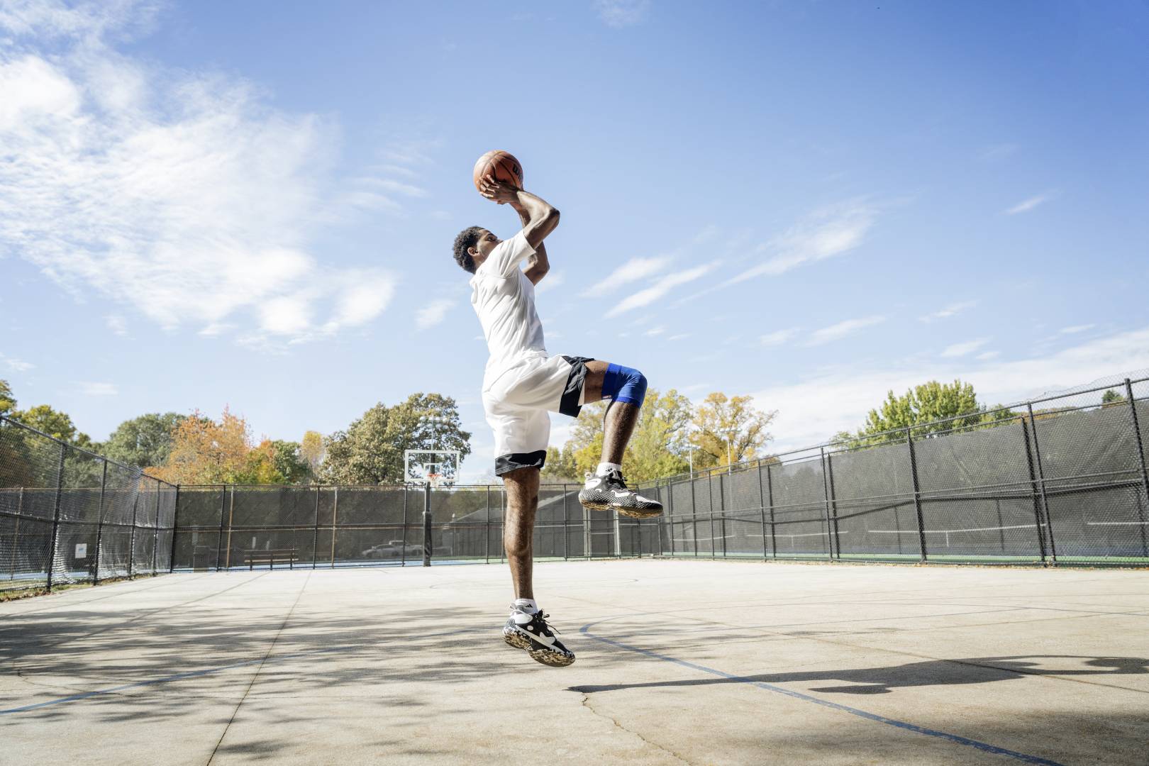 Basketballer springt und trägt Knie Bandage Basketballer springt auf einem Streetcourt für den Wurf ab	 Hände mit Ball überm Kopf	 ein Bein angewinkelt	 das andere senkrecht nach unten zeigend